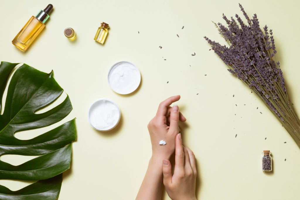 Top view and flat lay of woman holding cream on hands over white table with cosmetic products - avocado oil, cream and bamboo