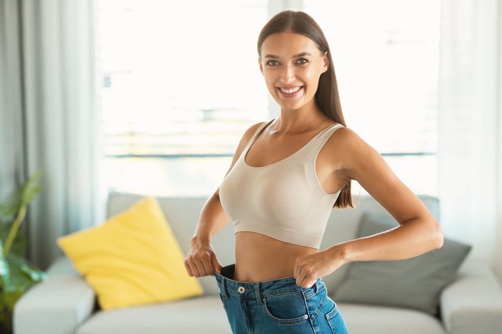 A woman looking in the mirror and applying SkinMedica® skincare products in Palm Springs Woman adjusting the waistband of loose-fitting pants after progress with medical weight loss.